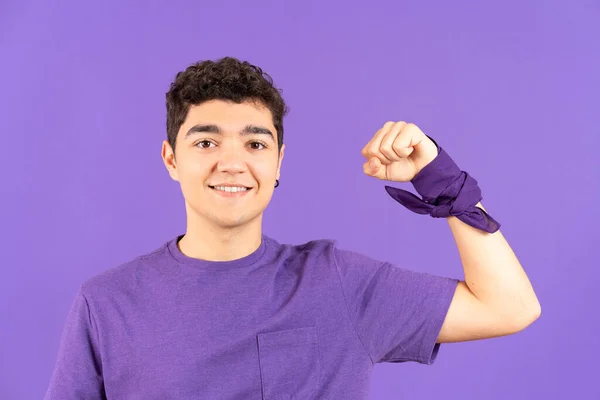 Front view of hispanic feminist teenager boy showing strength with raised arm isolated on purple background