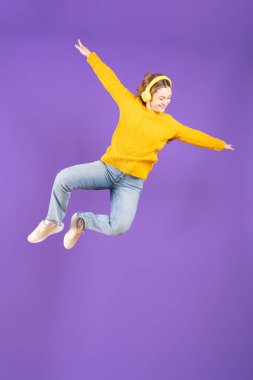 Pretty young woman dancing or levitating while listening to music on headphones isolated on purple background