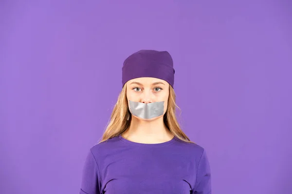 Young feminist woman covering mouth with adhesive tape isolated on purple background. Gender violence concept with copy space