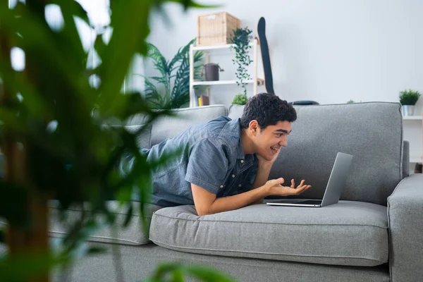 İspanyol genç çocuk kanepeye uzanıp dizüstü bilgisayarla konuşuyor. Ev konseptinden canlı yayın ve öğrenme