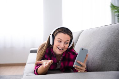 Pretty cheerful woman having video call on phone while lying down on couch and wearing headphones