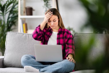 Young sad woman hand on head receiving bad news on laptop while sitting on couch in the living room at home