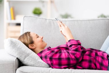 Young woman using phone while lying on back on sofa.