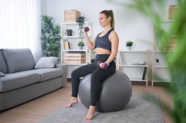 Young woman working out at home sitting on fitness ball and holding weights. Exercising and body care concept
