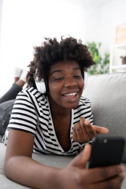 Kanepeye uzanmış gülümseyen Afro-Amerikan kadın video görüşmesi yapıyor ya da telefonla yayın yapıyor.