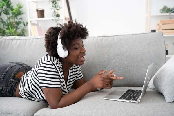 stock image African American young woman talking in front of laptop. Black woman broadcasting, live streaming or having a video call while lying on sofa at home. Online communication and social media concept