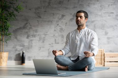 İspanyol yoga öğretmeni içeride dizüstü bilgisayarın önünde meditasyon yapıyor. Meditasyon dersini takip eden öğrenci bir paspasın üzerinde oturuyor.