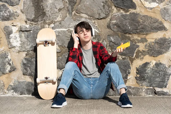 10代の少年は屋外でヘッドフォンで音楽を聴いています スケートボードの横の床に座ってリラックスした人 — ストック写真