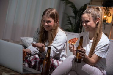 Pizza yerken görüntülü görüşme yapan iki genç kadın. Pijama giyen rahibeler kanepede yemek yiyor..