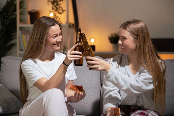 Two friends wearing pajamas toasting with beer at night. Sisters having dinner at home