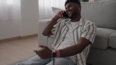 Afro-Amerikalı genç adam evde otururken telefonla konuşuyor..