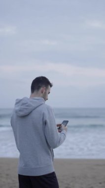 Sporcu sahilde selfie çekiyor. Genç adam deniz kenarında telefon kullanıyor.