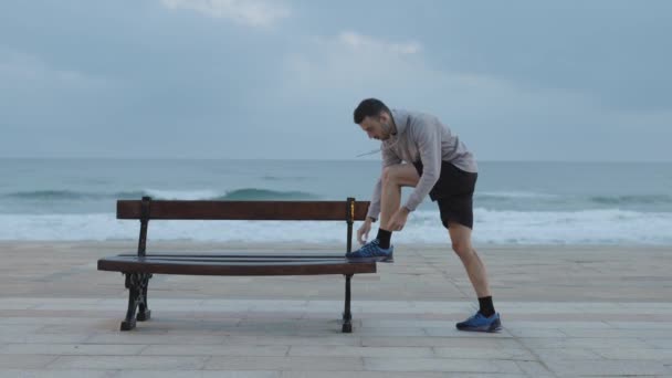 スポーツマントレーニング中にベンチで靴のレースを結ぶ 海岸の屋外で準備をするランナー — ストック動画