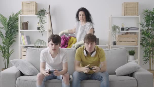 青少年男孩玩电子游戏 忽视母亲 女人喊着向儿子们扔衣服 平等和家务劳动分配概念 — 图库视频影像