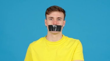 Genç adam mavi arka planda izole edilmiş ağzındaki bandı çıkarıyor. Konuşma ya da ifade özgürlüğü kavramı.