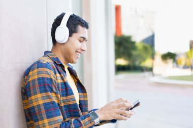 Müzik dinleyen ve kampüsün duvarına yaslanmış telefon kullanan mutlu bir öğrenci.