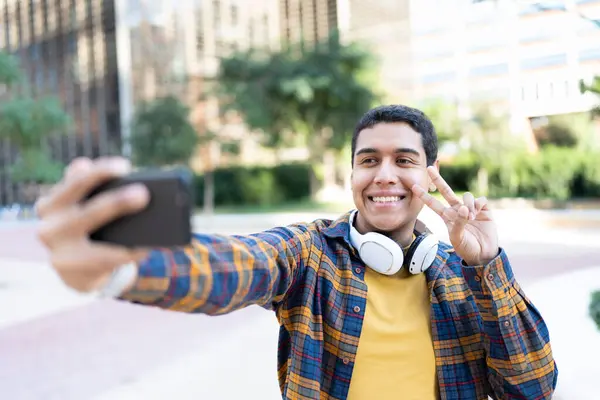 Smiling hispanic college student gesturing success while taking selfie outside the campus