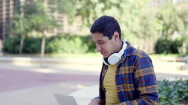 Yavaş çekimde yatay video. Üniversiteli bir Latin öğrenciden zum yaparak. Laptop kullanarak kampüsün dışında otururken başarıya işaret ediyor.