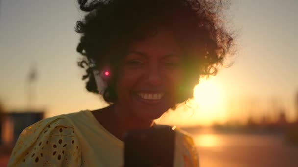 在日落时 一个笑容满面的非洲女人一边在户外用手机和耳机 一边笑 视频从她身上放大 — 图库视频影像