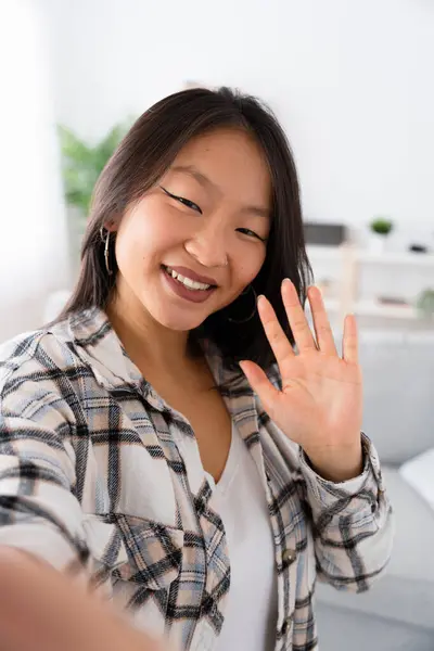 Vertical photo of a Chinese woman greeting at camera while taking selfie from home