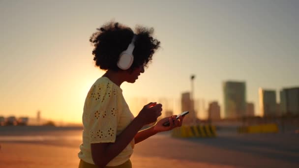 一个非洲女人在日落时用耳机和手机听音乐的视频 — 图库视频影像