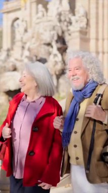 Güneşli bir günde iki yaşlı turistin güzel bir şehir parkını fıskiyeyle ziyaret ettiği dikey video.