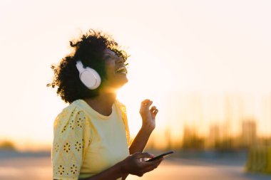 Gün batımında müzik dinleyen mutlu bir Afrikalı kadını aydınlatan güneş ışınları.