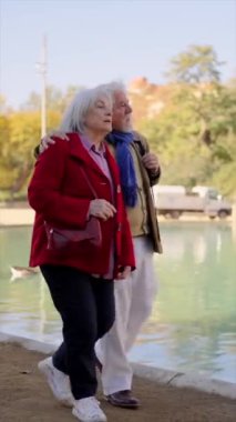 Şehir parkında yapay bir gölün kenarında birlikte yürüyen yaşlı bir çiftin dikey videosu.