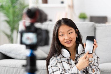 Çinli bir kadın kamerayla evde video dersi kaydediyor ve cep telefonunu gösteriyor.