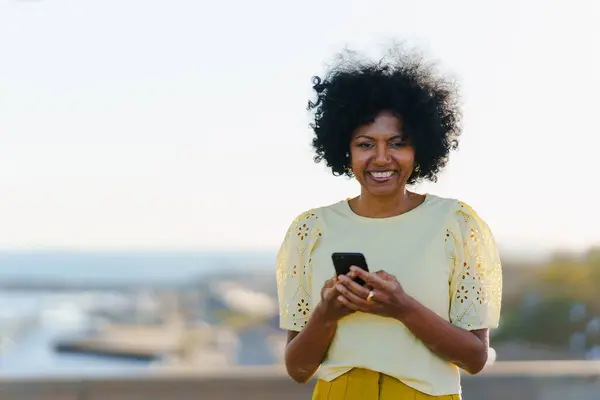 African woman smiling using phone outdoors standing in a city viewpoint during sunset