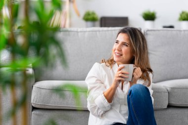 Dikkati dağılmış olgun bir kadın evde yerde oturmuş kahve içiyor.