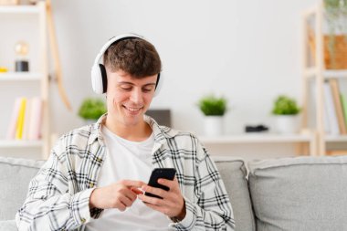 Genç çocuk oturma odasında telefonla müzik uygulaması kullanıyor. Şarkı seçen genç adam..