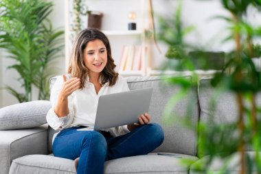 İnternetteki bir toplantıda dizüstü bilgisayar kullanan bir kadın evdeki kanepede oturuyor.