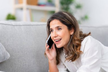 Kadın kanepede uzanıp evdeki cep telefonuyla konuşuyor.