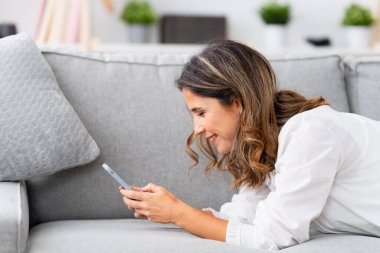 Mutlu, olgun bir kadının cep telefonuyla koltuğa uzanmış fotoğrafı.