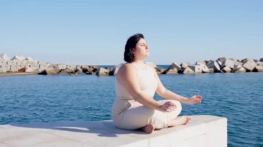 Kıvrımlı kadın deniz kenarında meditasyon yapıyor. Lotus pozundaki kişinin hareketli destansı videosu