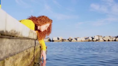 Kızıl saçlı genç kadın bir gölün suyuna eliyle dokunurken düşünüyor. Doğadaki düşünceli kadın.