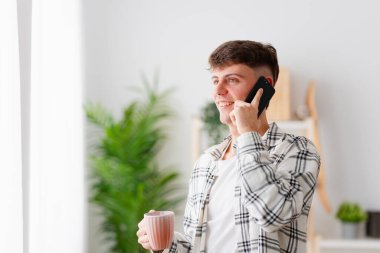 Genç ve gülümseyen bir adam kahve içiyor ve evdeki cep telefonuyla konuşuyor.