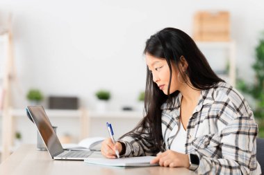 Çinli öğrenci not defteri ve dizüstü bilgisayar kullanarak evde oturuyor.