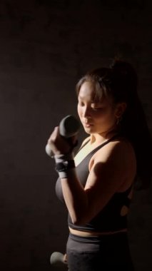 Genç Asyalı kadın karanlıkta ağırlık kaldırıyor. Sporcu kadının egzersiz yaptığı sinematik bir video. Kopya alanı ile spor motivasyonu ve kararlılık kavramı.