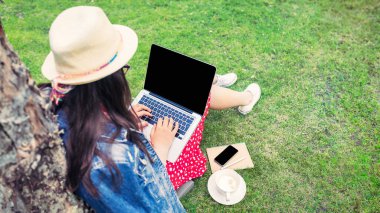 Genç bir kadın parkta çimlerde otururken boş ekranlı dizüstü bilgisayar kullanıyor.