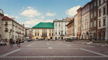 Krakow, Polonya - 29 Temmuz 2024; Polonya 'nın Krakow kentindeki Küçük Pazar Meydanı' nın sakin manzarası, tarihi renkli binalar, bir kaldırım taşı plaza ve güneşli bir günde gezinen insanlar ve mavi bir gökyüzü ve dağınık bulutlar..