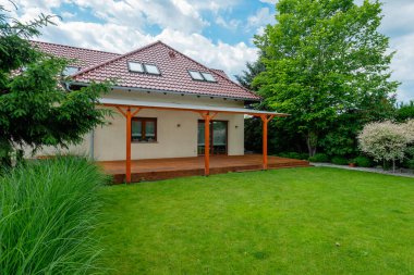 Kırmızı kiremit çatılı modern banliyö evi, şeffaf pergola ile ahşap güverte, ve güneşli bir günde yeşil çimenler ve yemyeşil ağaçlar içeren güzel manzaralı arka bahçe.