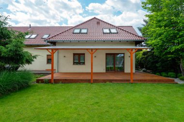 Kırmızı kiremit çatılı modern banliyö evi, şeffaf pergola ile ahşap güverte, ve güneşli bir günde yeşil çimenler ve yemyeşil ağaçlar içeren güzel manzaralı arka bahçe.