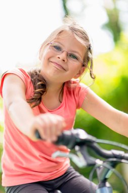 Yeşil bir parkta bisiklet süren gözlüklü genç bir kız aktif bir yaşam tarzı ve yaz tatilinin tadını çıkarıyor.