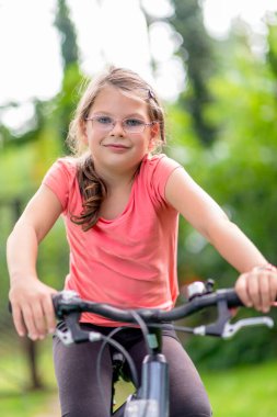 Yeşil bir parkta bisiklet süren gözlüklü genç bir kız aktif bir yaşam tarzı ve yaz tatilinin tadını çıkarıyor.