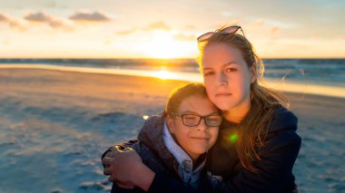 Kardeşler gün batımında plajda kucaklaşıyorlar, tatilde huzurlu bir anın tadını çıkarıyorlar, arka planda sıcak altın ışık ve okyanus var.