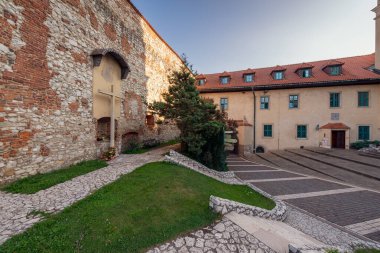 Tyniec, Polonya - 29 Temmuz 2024; Ortaçağ taş ve tuğla duvarlı Tyniec Manastırı tarihi avlusu, büyük ahşap haç, yeşil pencereler, kırmızı çatı ve güneşli bir günde sakin bahçe yolu