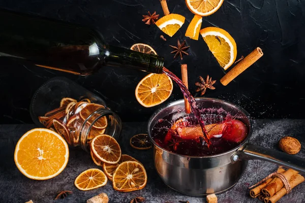 Falling mulled wine ingredients, citrus fruits, cinnamon and star anise fly from a height and create splashes. A cauldron of mulled wine into which fruit and spices are falling and wine pouring. In the dark background are dried citrus fruits.