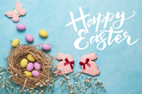 Banner for the Easter holiday. Colored eggs in a straw nest and white spring flowers on a blue background. Place for text. Top view.